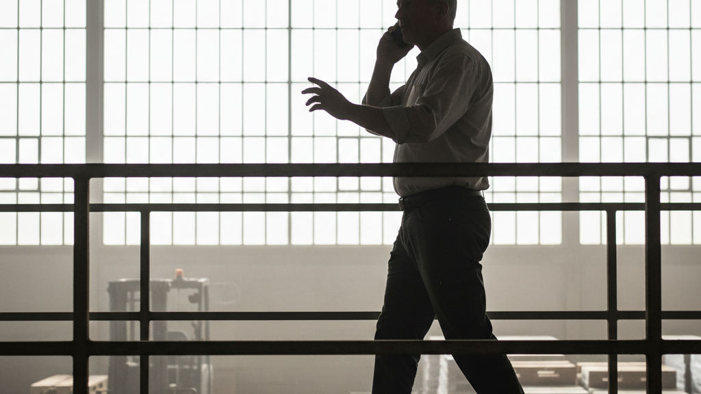 Professional in a leather jacket uses a phone for logistics inteligente while walking in a sunlit warehouse